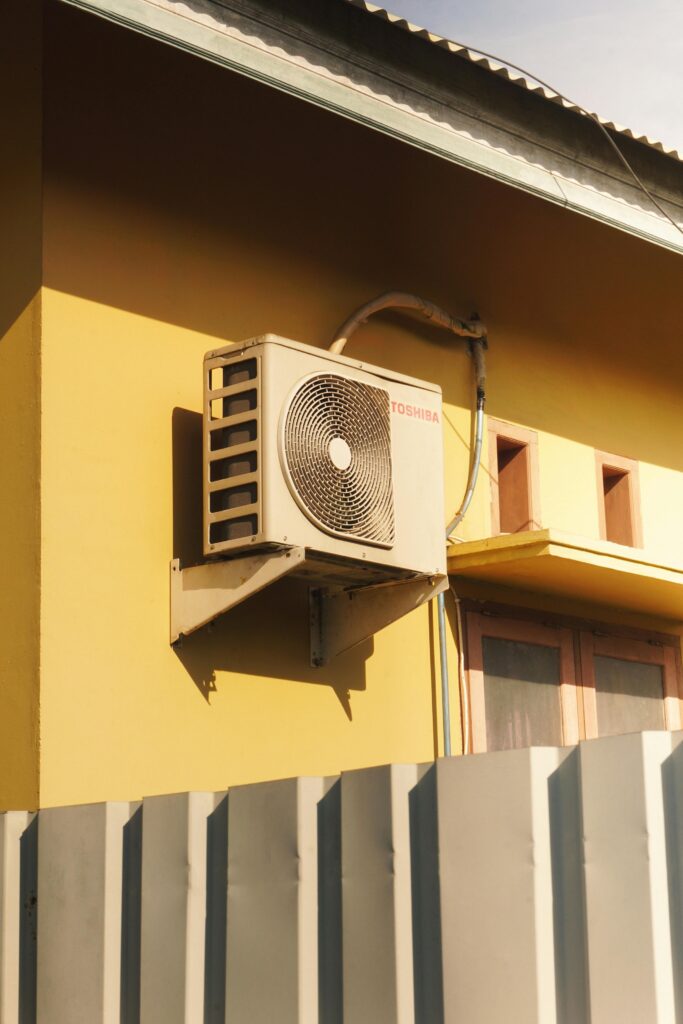 pexels-photo-14086132-14086132 Outdoor air conditioner unit attached to a sunny yellow building facade.